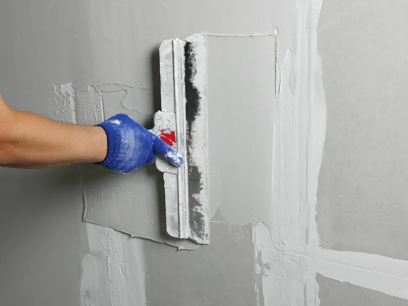 A gloved hand drawing a wide knife of compound across a patched drywall wall