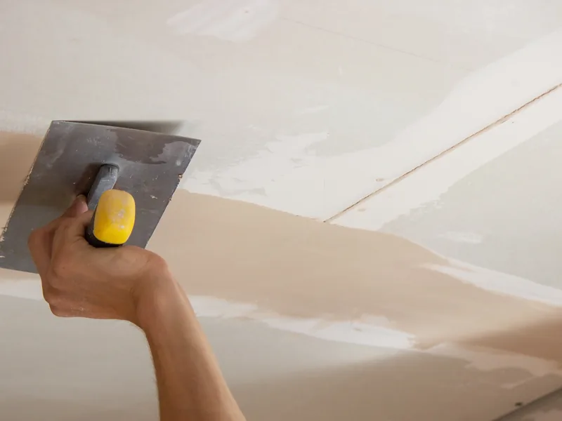 A trowel drawing joint compound along the drywall join between wall and ceiling