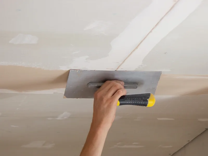 A hand flattening compound across an overhead ceiling seam, the drywall finishing included in every repair price