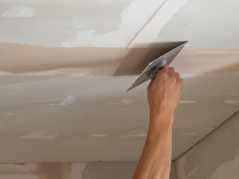 A trowel worked along a ceiling seam from below at arm's length, the drywall finishing a price covers