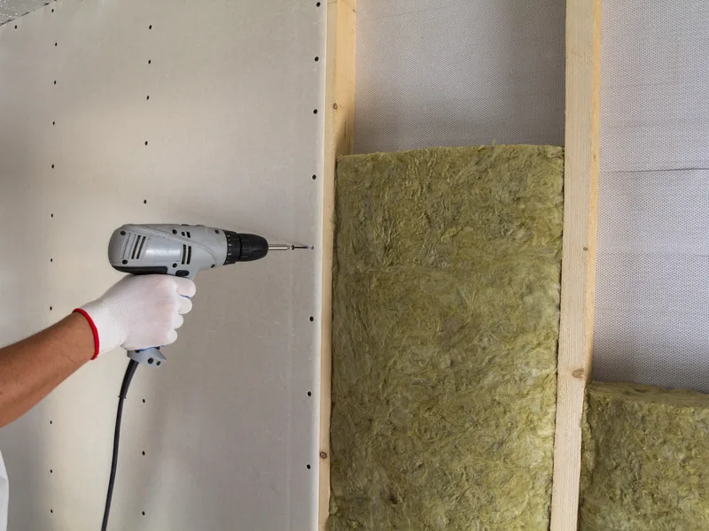 A screw gun driving a drywall fastener through board beside an open insulated bay, before the wall is closed