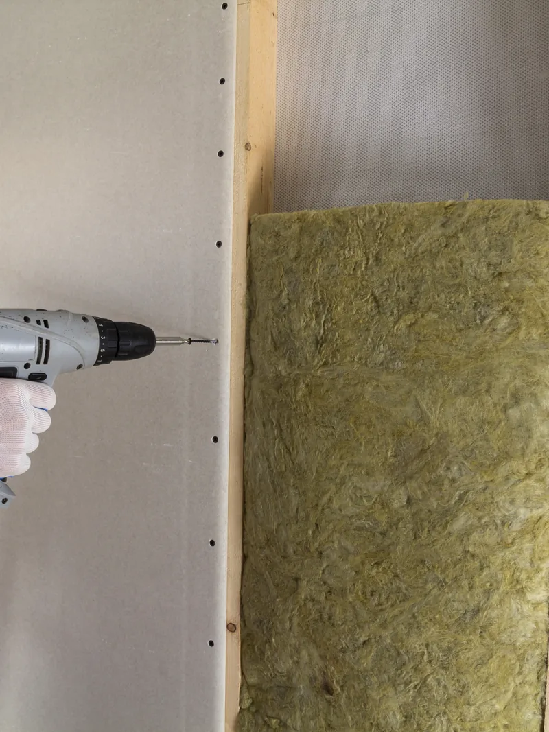 A screw gun driving a fastener through board beside an open insulated bay