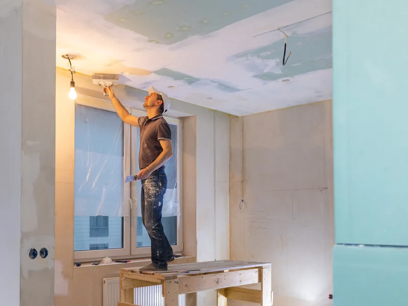 A worker standing on a box to run drywall compound along the top of a wall