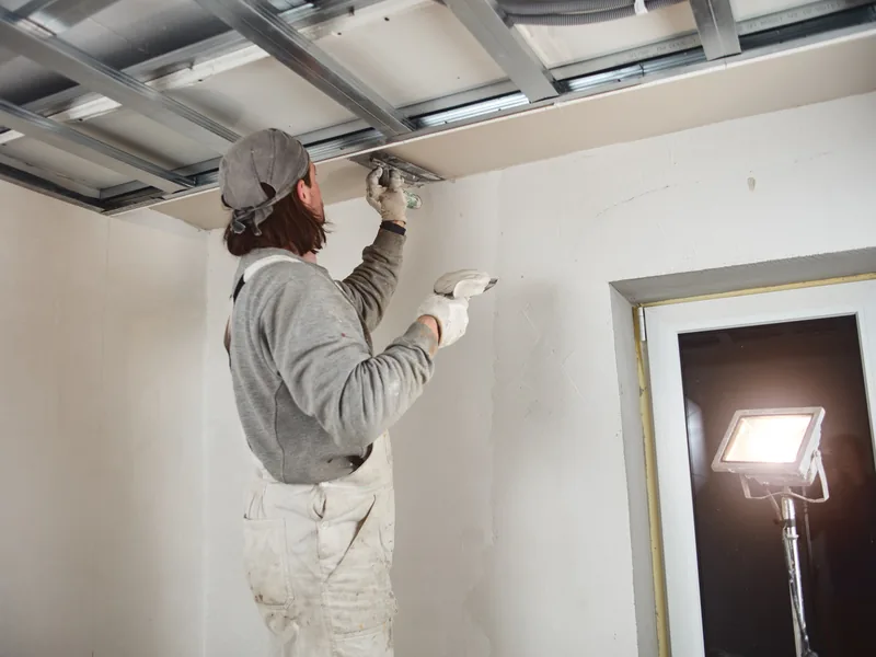 A worker finishing a wall beside a work light angled across the surface