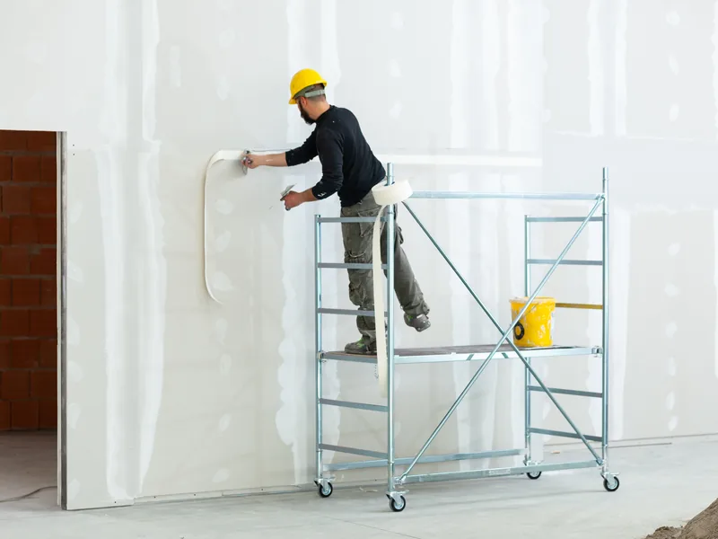 A worker on a rolling tower taping the upper part of a tall wall in an office interior