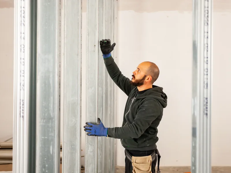 A worker setting a steel stud into a track inside a part-framed opening, dividing a working building