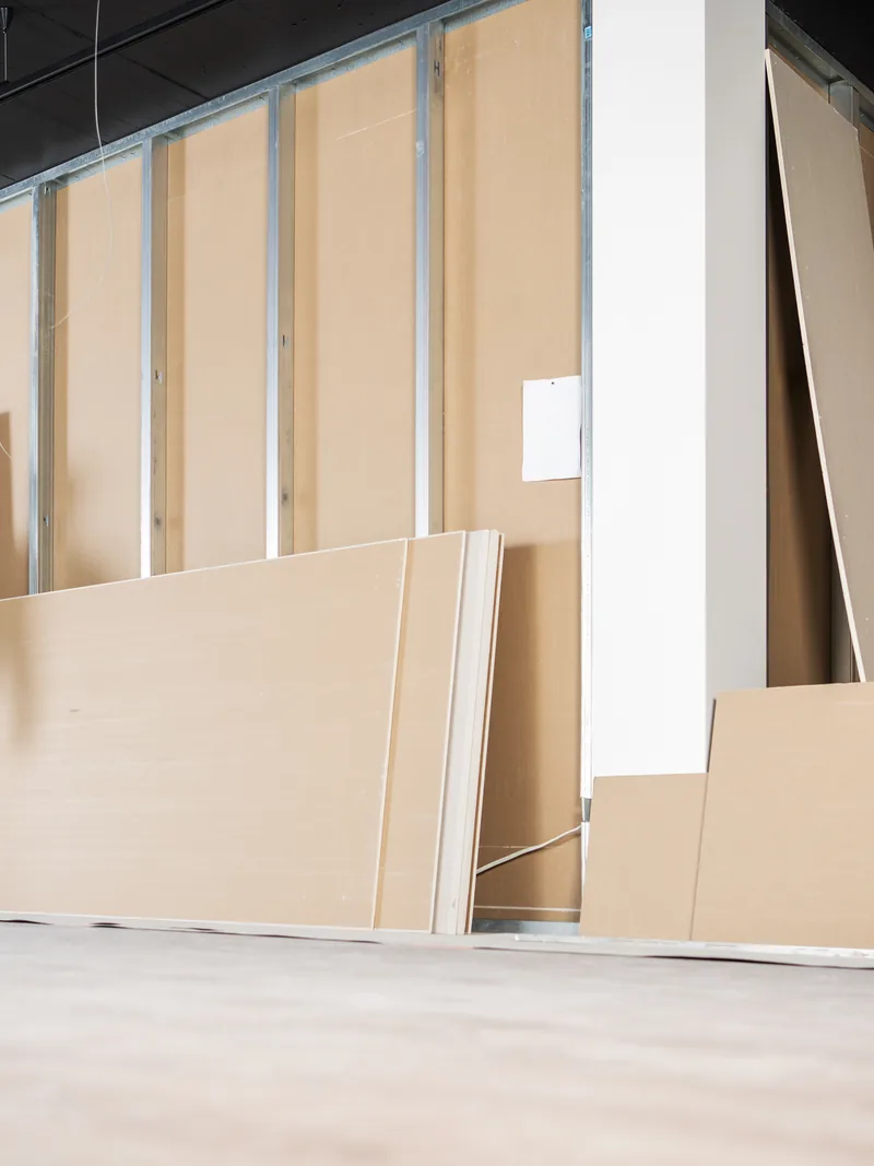 A worker steadying a drywall panel against steel studs in a long commercial space