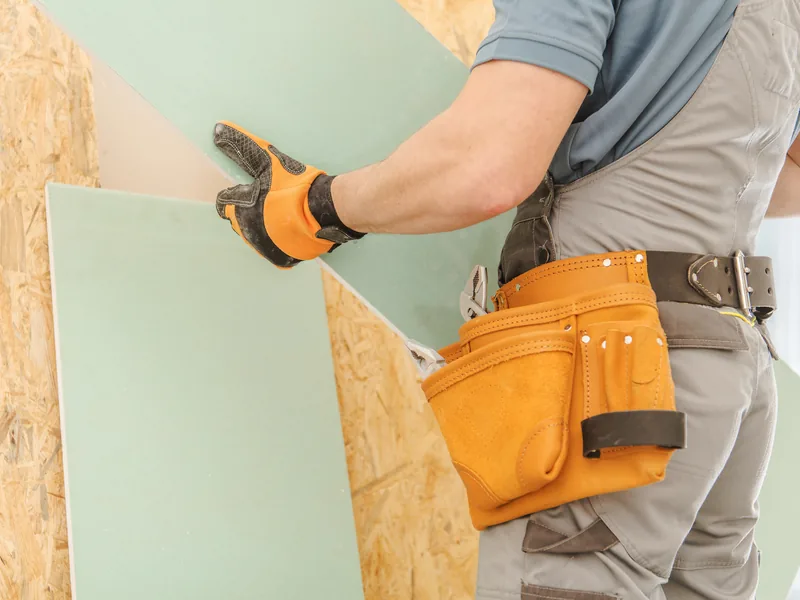 Gloved hands lowering a green moisture-resistant drywall panel onto a sheathed wall
