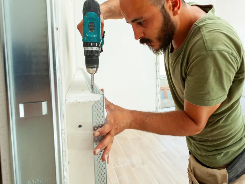 A worker fixing a metal corner bead to an outside corner with a drill, a repair that stops short of replacement