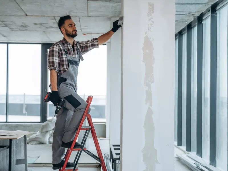 A worker on a stepladder finishing a wall beside a glazed office partition while the business stays open