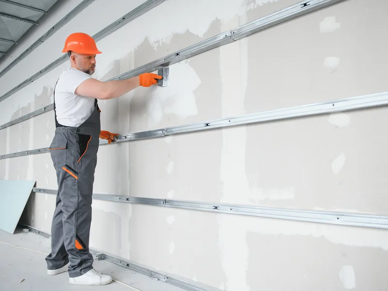 A worker running a drywall knife along a wall lined with horizontal steel furring