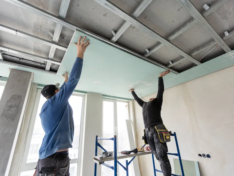 Two workers fitting panels into an exposed ceiling grid from step platforms in a commercial interior