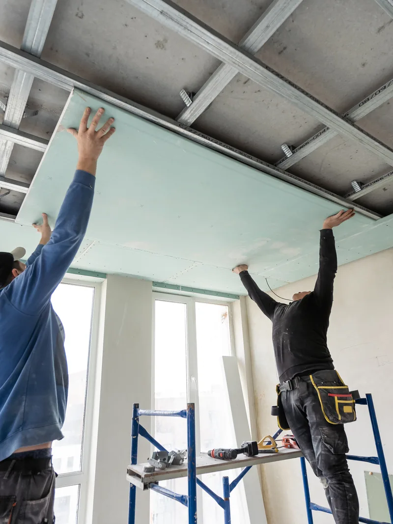 Two workers fitting panels overhead in a commercial space from step platforms