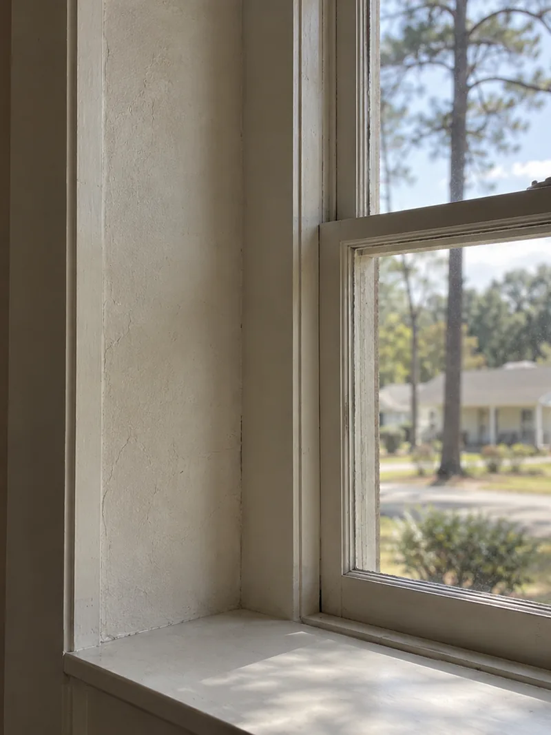 A window set in a deep wall reveal with its original casing, looking out to a front lawn