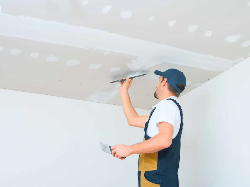 A worker finishing the drywall corner where a new ceiling meets the wall