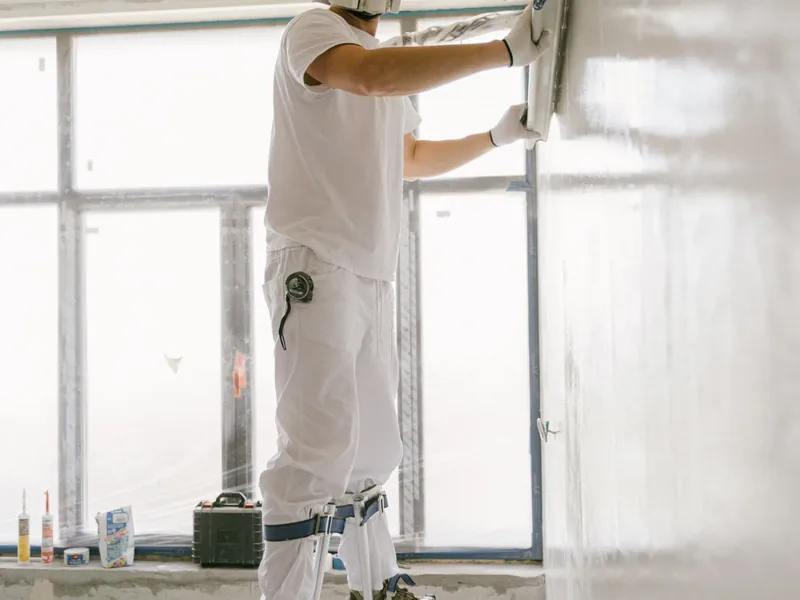 A worker skim-coating the top of a drywall wall from a stepladder