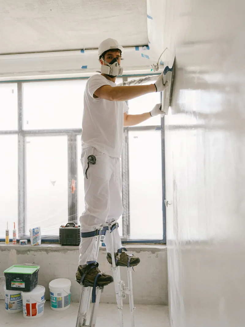 A worker skim-coating the top of a wall from a stepladder as part of the finish