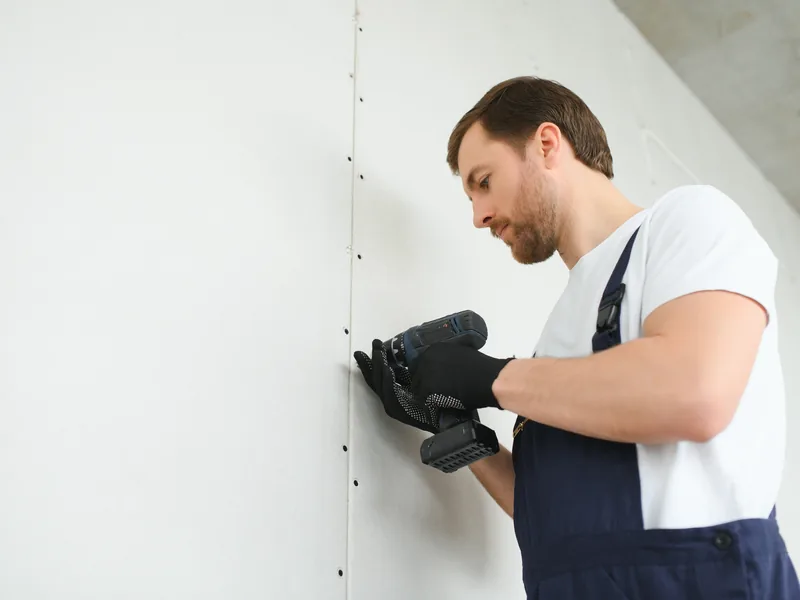 A worker running a rasp along the cut edge of a drywall panel held against the wall