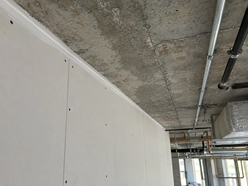 The top of a boarded wall sealed to the underside of a concrete slab in an industrial interior, services running across the soffit beyond