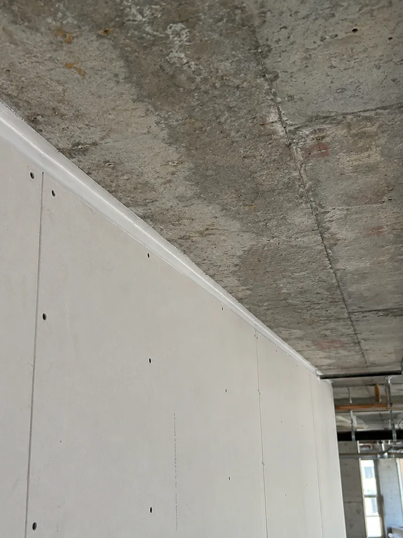 The top of a boarded wall sealed to the underside of a concrete slab, with services running across the soffit beyond