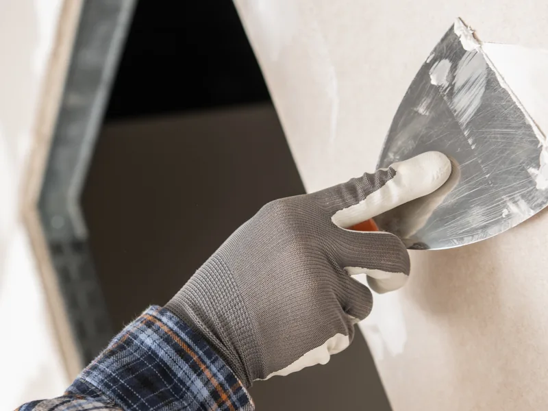 A gloved hand carrying a trowel of drywall compound to a dark wall opening