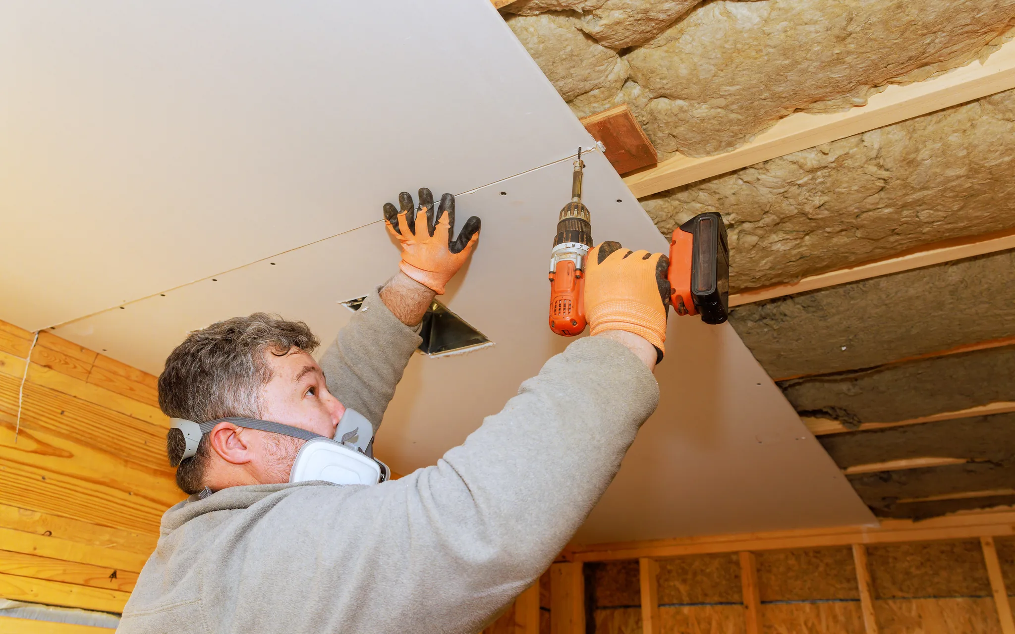 A worker installing ceiling drywall, driving screws into a panel between insulated joists