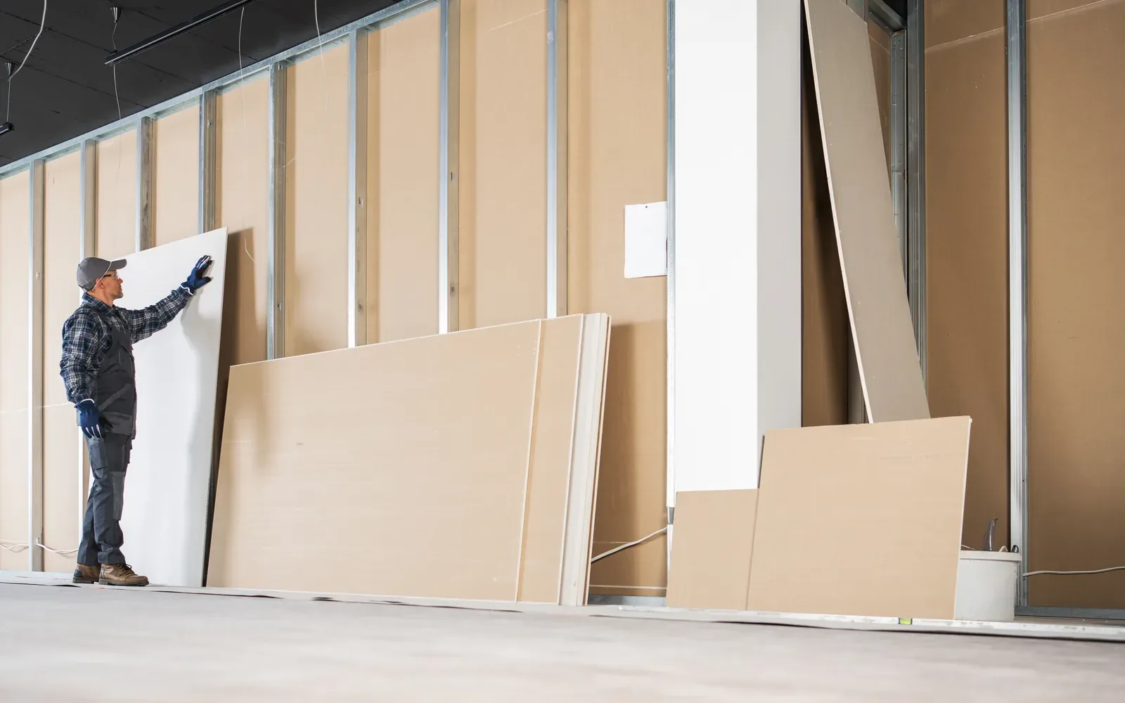 A worker steadying a drywall panel against steel studs in a long commercial space