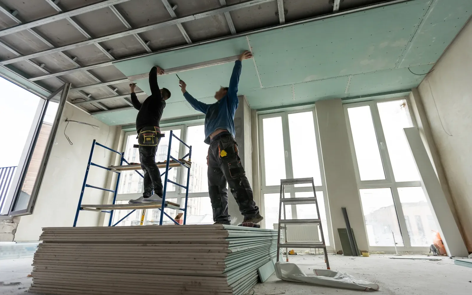 Two workers on a platform lifting a green moisture-resistant drywall panel to a ceiling frame