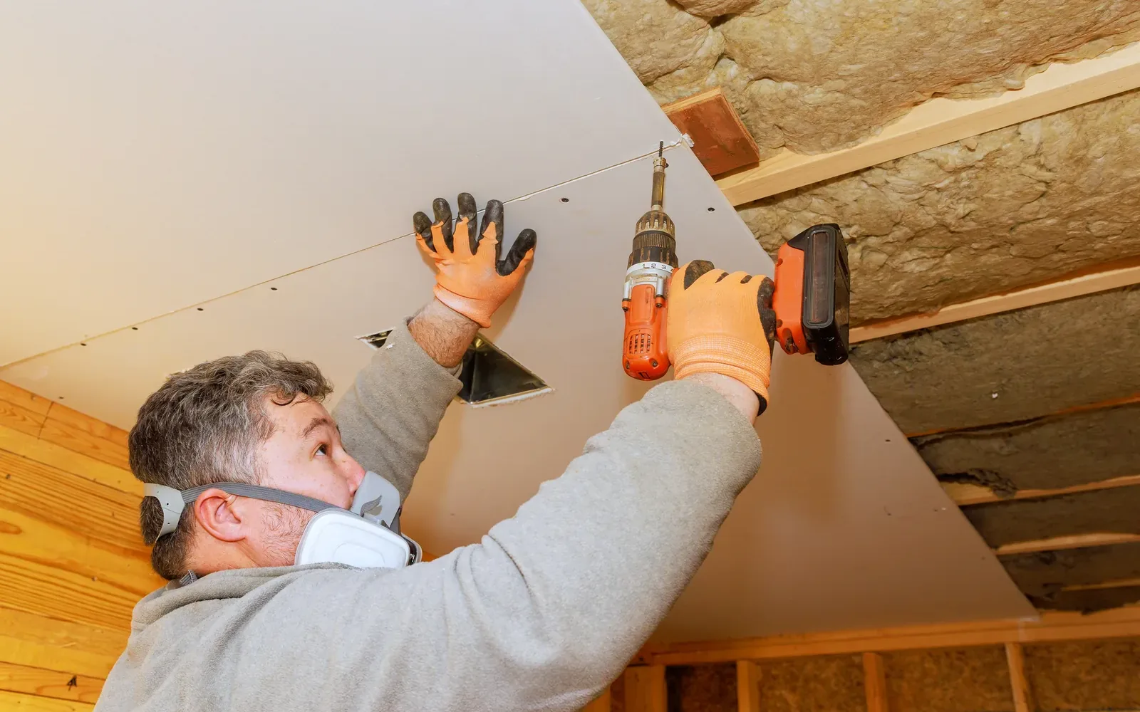 Ceiling drywall going in, a worker driving screws into a panel between insulated joists