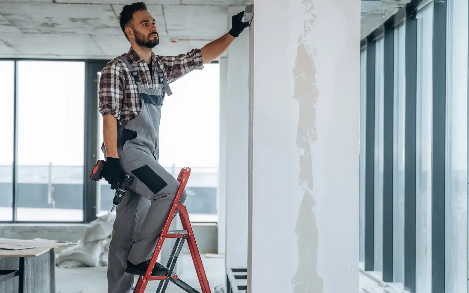 A worker on a stepladder finishing a drywall wall beside a glazed office partition