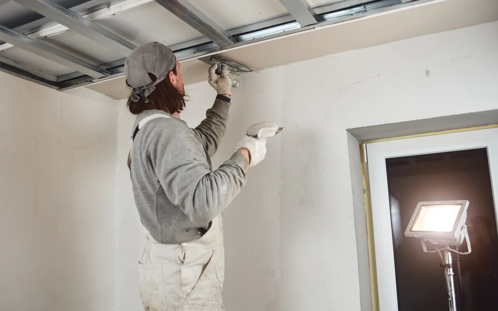 A worker finishing a drywall wall beside a work light angled across the surface to read the texture