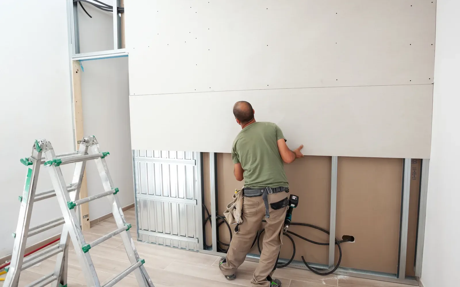 A worker boarding out a drywall wall with a stepladder and offcuts beside him