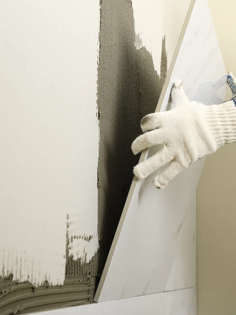 Gloved hands pressing tile onto a combed bed of adhesive over a finished wall