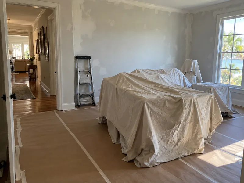One room of a lived-in house masked for painting, furniture sheeted in the middle, the hallway beyond untouched