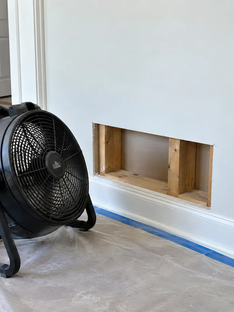 A floor fan drying an opened wall bay of clean studs behind a taped dust sheet