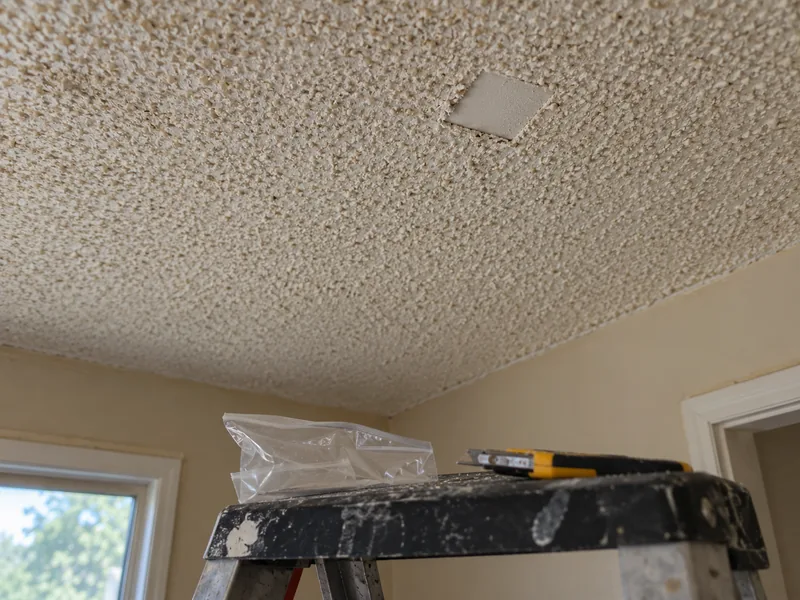 A small square cut out of an aged popcorn ceiling with a specimen bag and knife on a stepladder below, sampled before anything is disturbed