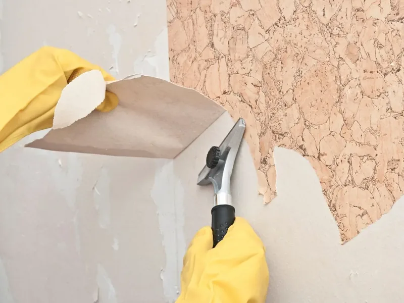 Gloved hands working a scraper under a sheet of loosened wallpaper