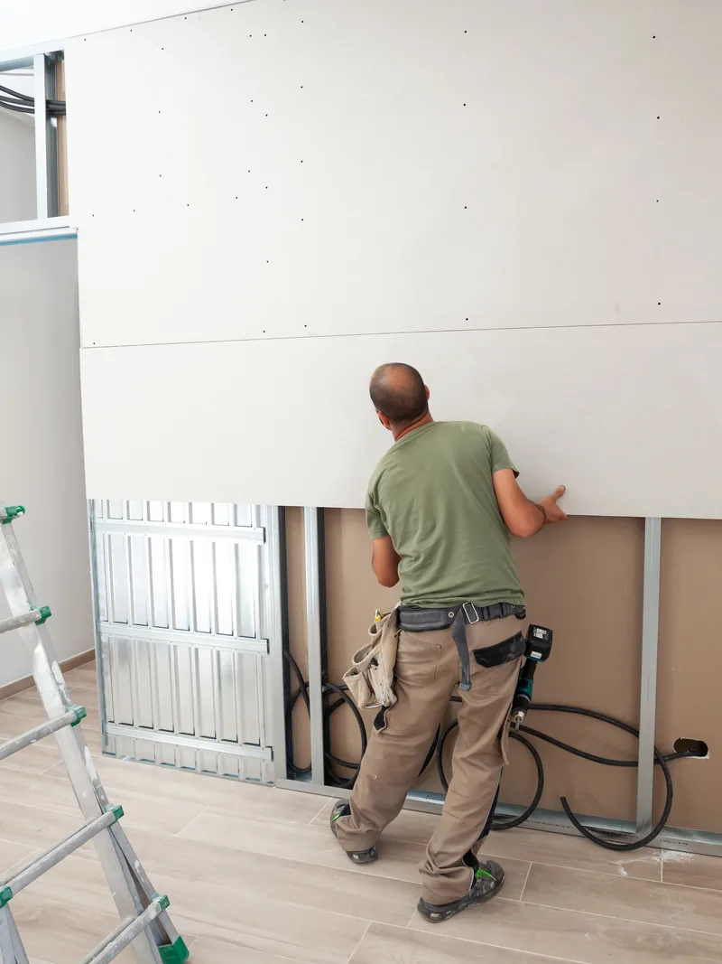 A worker boarding out a wall with offcuts and a stepladder beside him